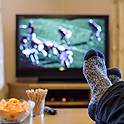 In the background a TV with a football game. In the foreground feet on a table with a bowl of snack to the left. 