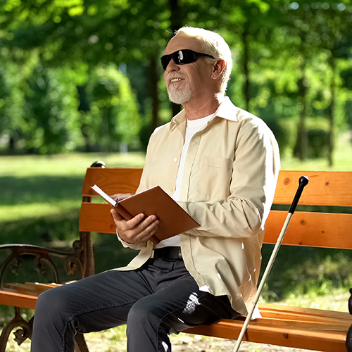 A man with sunglasses on and a cane sitting next to him sitting on a bench outside reading a Braille book