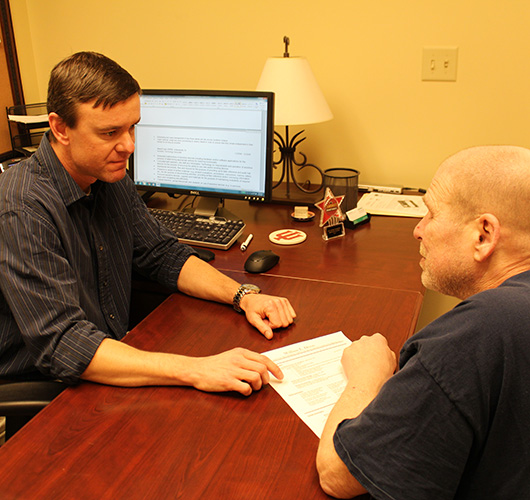 A man sits across a desk from another man they are talking a reviewing paperwork.