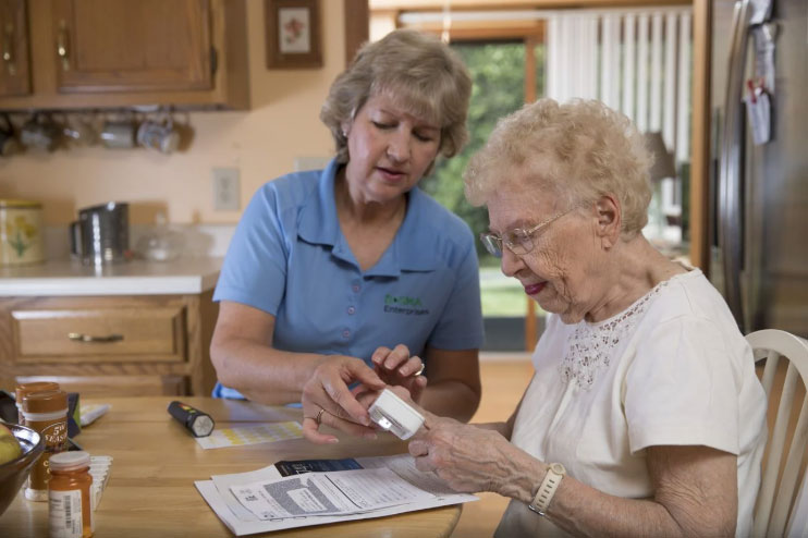 Two women sitting at table while one helps the other use a magifying device to review instructions.