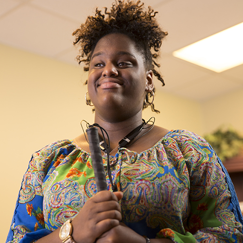 A woman holding her white cane with both hands in front of her and smiling.