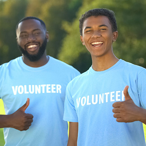 Two men wearing volunteer shirts look at the camera, smiling and giving a thumbs up.