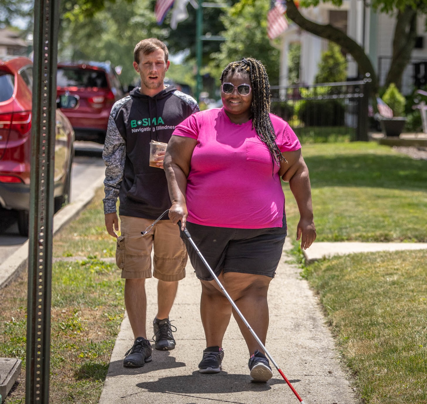 A woman walking on a sidewalk with a white cane and a man is walking just behind her. 