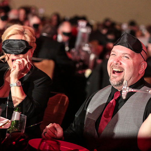 A man and a woman dressed nicely at a table with sleep shades on and laughing. 