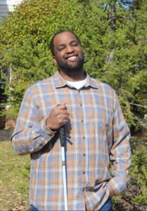 Ory Watson standing in a park smiling at the camera holding a white cane.