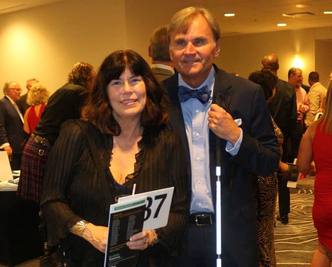 Man with white cane and woman with auction placard smiling at camera.