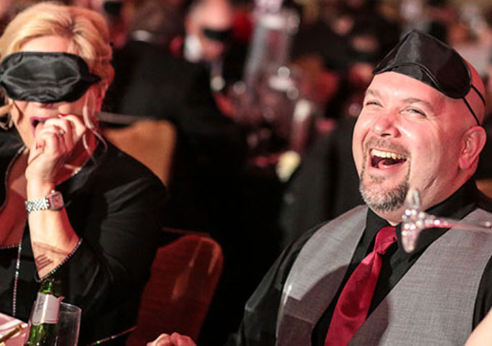 A woman with a sleep shade on and a man with a sleep shade pulled up to his forehead laugh while at the Dining in the Dark event.