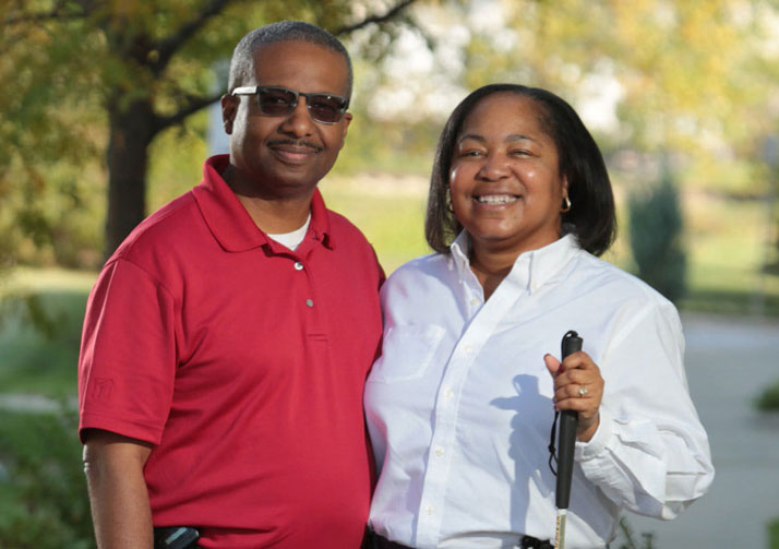 Man with woman holding white cane standing outside smiling at camera.