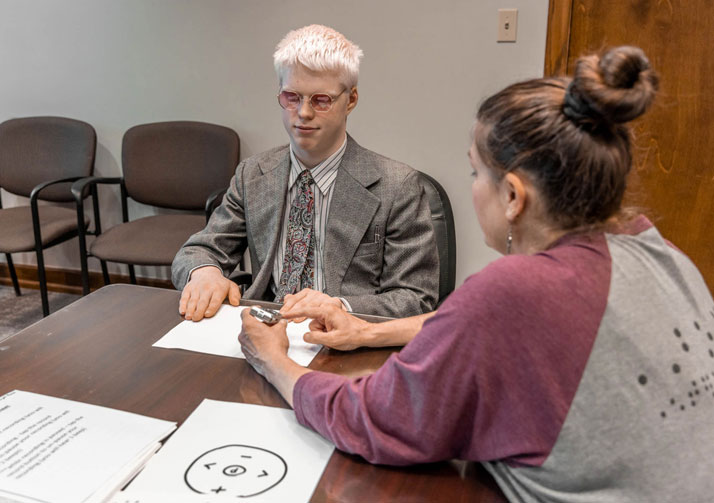 Man and woman sitting at a table while the woman trains man on an assistive device.