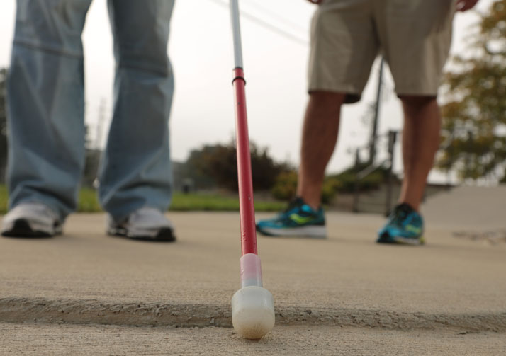 Close up picture of the end of a white cane on the sidewalk with two pairs of legs visible in the background.